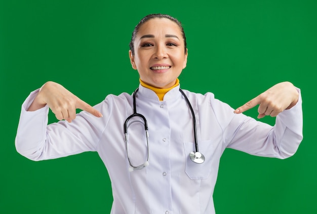 Medico della giovane donna in camice bianco della medicina con lo stetoscopio intorno al collo che sembra sorridente sicuro che indica con le dita se stessa che sta sopra la parete verde