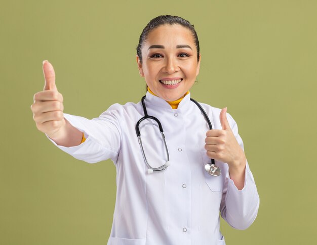 Medico della giovane donna in camice bianco della medicina con lo stetoscopio intorno al collo che sorride fiducioso che mostra i pollici su che stanno sopra la parete verde