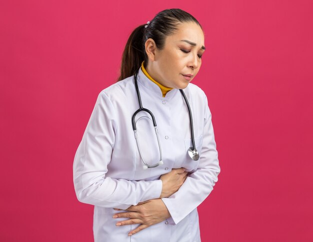 Medico della giovane donna in camice bianco della medicina con lo stetoscopio intorno al collo che tocca la sua pancia che sembra malessere che sente dolore che sta sopra la parete rosa