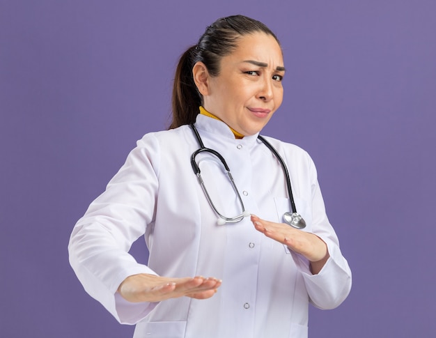Medico della giovane donna in camice bianco della medicina con lo stetoscopio intorno al collo con l'espressione disgustata che fa il gesto di difesa con le mani che stanno sopra la parete viola