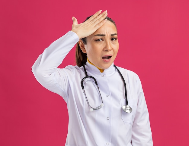 Medico della giovane donna in camice bianco della medicina con lo stetoscopio intorno al collo confuso con la mano sulla sua testa per errore che sta sopra la parete rosa