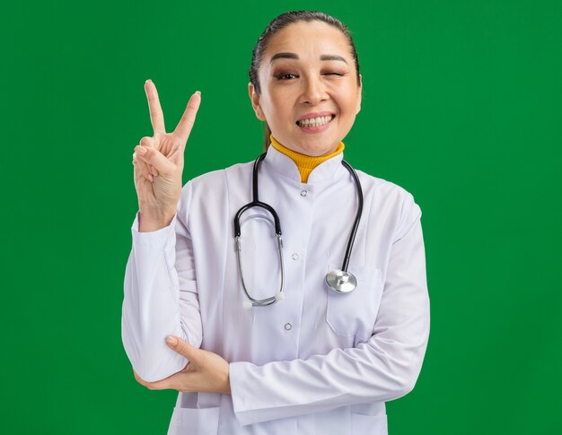 Medico della giovane donna in camice bianco della medicina con lo stetoscopio intorno al collo felice e gioioso che strizza l'occhio e che sorride mostrando il v-sign che sta sopra la parete verde