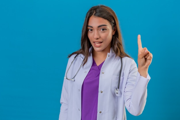 Medico della giovane donna in camice con il dito indicante sicuro e felice sorridente del fonendoscopio su sopra fondo blu isolato