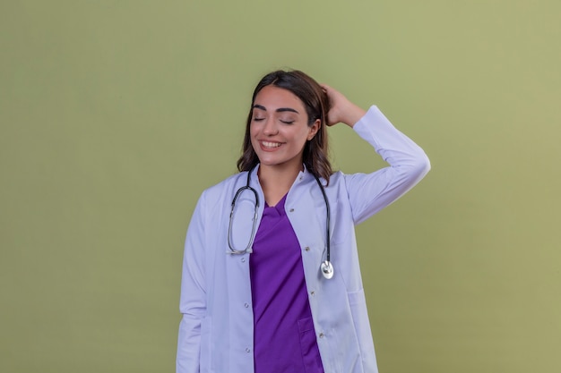 Medico della giovane donna in camice con phonendoscope che sembra sicuro con il sorriso sul fronte e sugli occhi chiusi che toccano capelli che controllano fondo verde