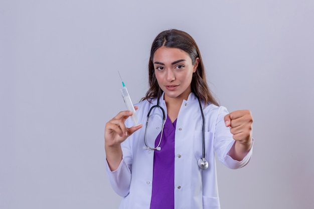 Medico della giovane donna in camice con phonendoscope che sembra siringa sicura della tenuta e che mostra pugno alla macchina fotografica sopra fondo bianco isolato