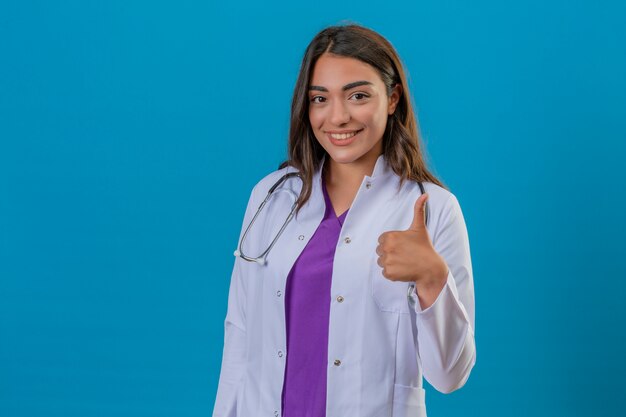 Medico della giovane donna in camice con phonendoscope che sta con il grande pollice di sorriso su sopra fondo blu isolato