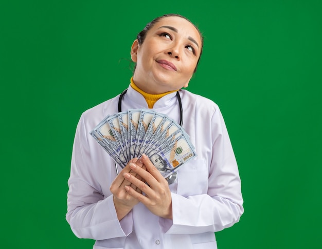 Medico della giovane donna in camice medico bianco con lo stetoscopio intorno al collo che guarda felice e compiaciuto che sta sopra la parete verde