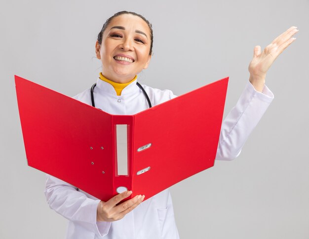 Medico della giovane donna in camice medico bianco con lo stetoscopio intorno al collo che tiene cartella rossa felice ed eccitata