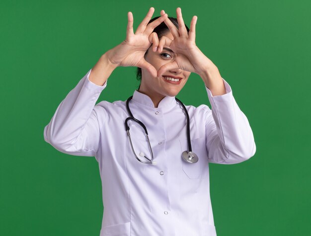 Medico della giovane donna in cappotto medico con lo stetoscopio guardando la parte anteriore con il sorriso sul viso che fa il gesto del cuore con le dita in piedi sopra la parete verde