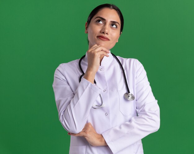 Medico della giovane donna in cappotto medico con lo stetoscopio intorno al collo che osserva in su con espressione pensierosa che pensa che si leva in piedi sopra la parete verde