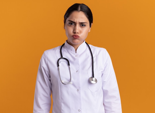 Medico della giovane donna in cappotto medico con uno stetoscopio intorno al collo guardando le guance di soffiaggio anteriore con espressione infastidita in piedi sopra la parete arancione