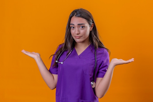 Medico della giovane donna in uniforme medica con il fonendoscopio che sta con l'espressione confusa con le armi e le mani sollevate sopra il fondo arancio isolato