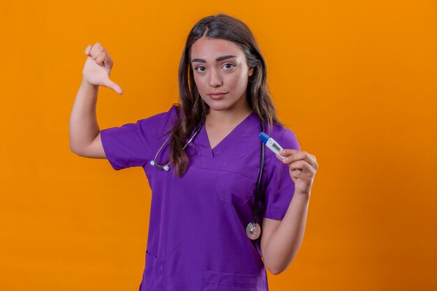 Medico della giovane donna in uniforme medica con il pollice infelice del termometro della tenuta del fronte dello stetoscopio giù che mostra antipatia sopra fondo arancio isolato