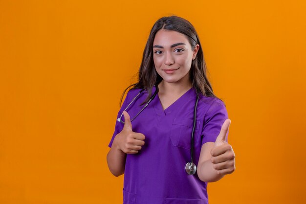 Medico della giovane donna in uniforme medica con phonendoscope che sta con il sorriso sul fronte che mostra i pollici su sopra fondo arancio isolato