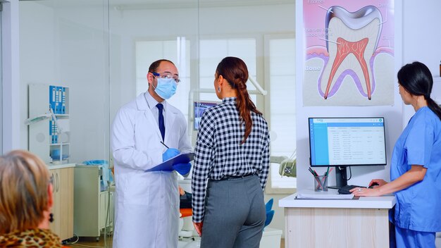 Medico dentista che interroga la donna e prende appunti sugli appunti in piedi nell'area di attesa. Giovane paziente che spiega il problema dentale allo stomatologo che parla nella reception affollata della clinica.