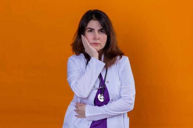 Medico di mezza età che indossa camice bianco e con lo stetoscopio scettico e nervoso disapprovazione espressione sul viso con la mano sulla guancia sul muro arancione