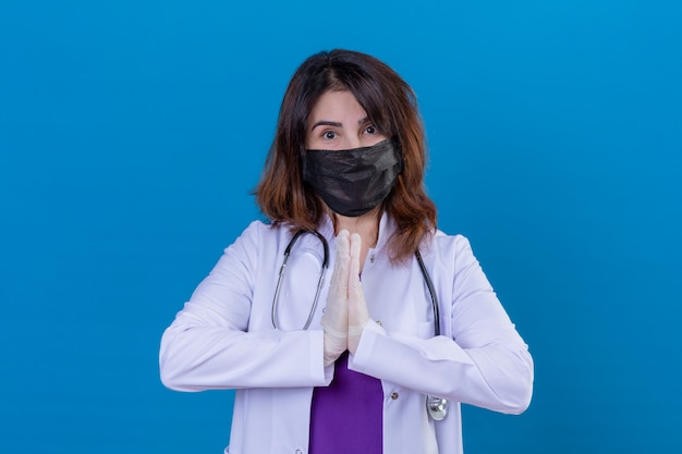 Medico di mezza età che indossa camice bianco in maschera protettiva nera e con lo stetoscopio si tengono per mano nel gesto del namaste di preghiera