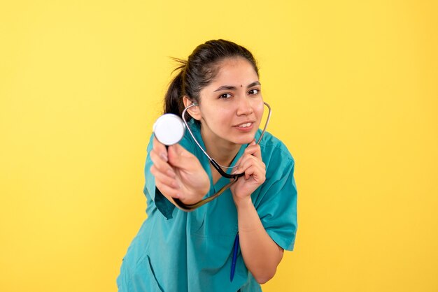 Medico donna beata vista frontale in uniforme che mostra lo stetoscopio su sfondo giallo