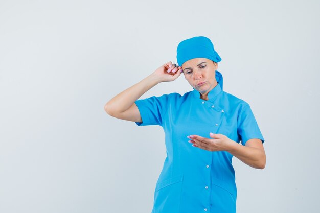 Medico donna che finge di guardare il telefono cellulare in uniforme blu e guardando pensieroso, vista frontale.