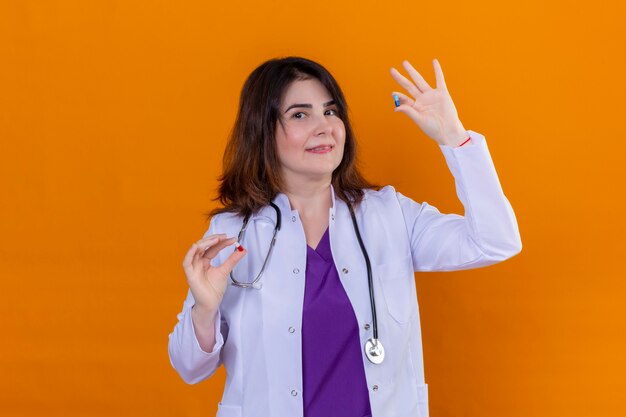 Medico donna di mezza età che indossa camice bianco e con lo stetoscopio tenendo le pillole in mano guardando la fotocamera sorridente in piedi su sfondo arancione