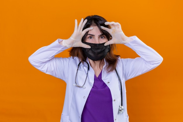 Medico donna di mezza età che indossa camice bianco nella maschera facciale protettiva nera e con lo stetoscopio cercando di aprire gli occhi con le dita assonnate e stanche per la stanchezza mattutina in piedi su te isolato