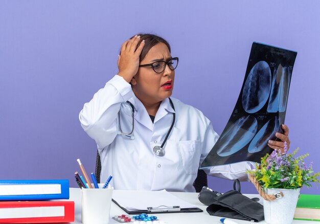 Medico donna di mezza età in camice bianco con stetoscopio che tiene raggi x guardandolo con espressione confusa con la mano sulla testa seduta al tavolo sul muro blu