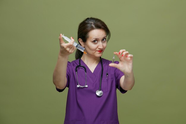 Medico donna di mezza età incerto che indossa uniforme e stetoscopio intorno al collo guardando la telecamera che allunga la siringa con ago e fiala medica verso la telecamera isolata su sfondo verde oliva