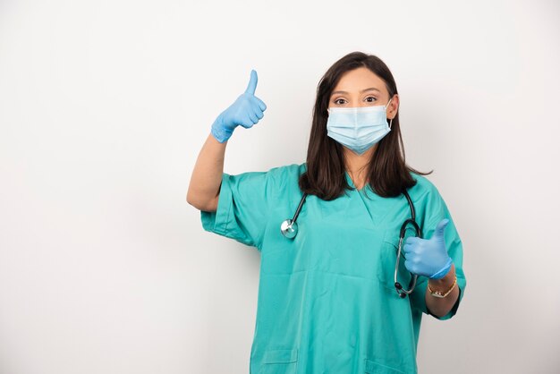 Medico donna in maschera che mostra i pollici in su su sfondo bianco. Foto di alta qualità