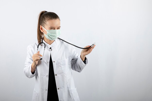 Medico donna in uniforme bianca che indossa una maschera medica e tiene in mano uno stetoscopio