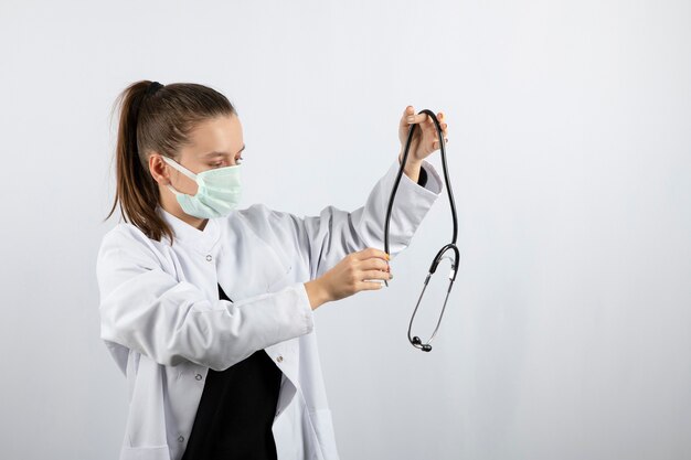 Medico donna in uniforme bianca che indossa una maschera medica e tiene in mano uno stetoscopio