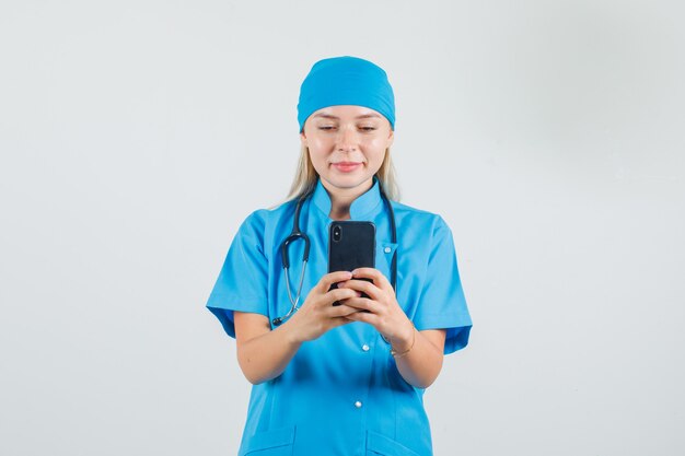 Medico donna sorridente durante l'utilizzo di smartphone in uniforme blu e guardando occupato