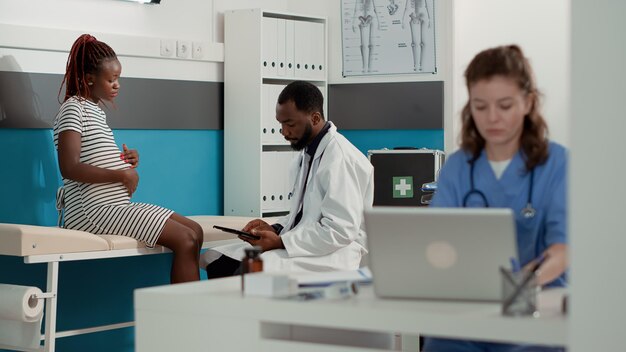 Medico e donna afroamericani che fanno consulenza sulla gravidanza in ufficio presso la clinica sanitaria. Specialista della maternità che prende appunti sulla tavoletta digitale durante la visita di controllo con una paziente incinta.