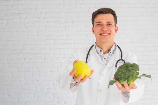 Medico felice maschio sorridente che offre mela e broccoli