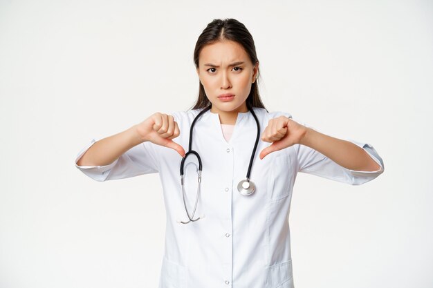 Medico femminile asiatico dall'aspetto serio deluso, medico ospedaliero mostra i pollici verso il basso con disapprovazione, antipatia per smth cattivo, in piedi su sfondo bianco.