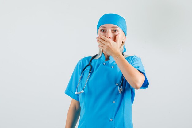 Medico femminile che copre la bocca con la mano in uniforme blu e sembra smemorato