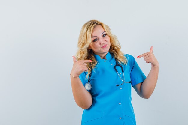 Medico femminile che indica a se stessa in uniforme blu e che sembra fiducioso.
