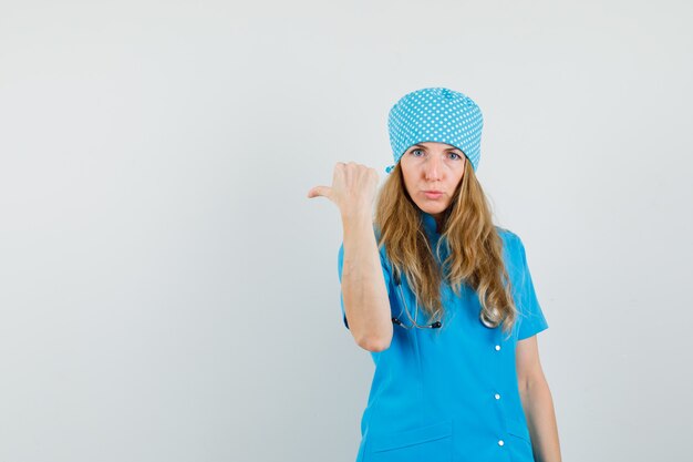 Medico femminile che indica il pollice fino a lato in uniforme blu e che sembra serio.