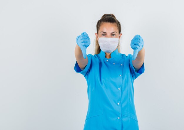 Medico femminile che mostra i pollici giù in uniforme blu, maschera, guanti e sguardo scontento.