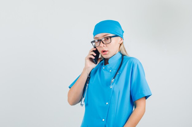 Medico femminile che parla sullo smartphone in uniforme blu, occhiali e che sembra impotente.