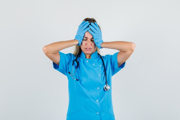 Medico femminile che tiene le mani sulla testa in uniforme blu, guanti e che sembra dispiaciuto.