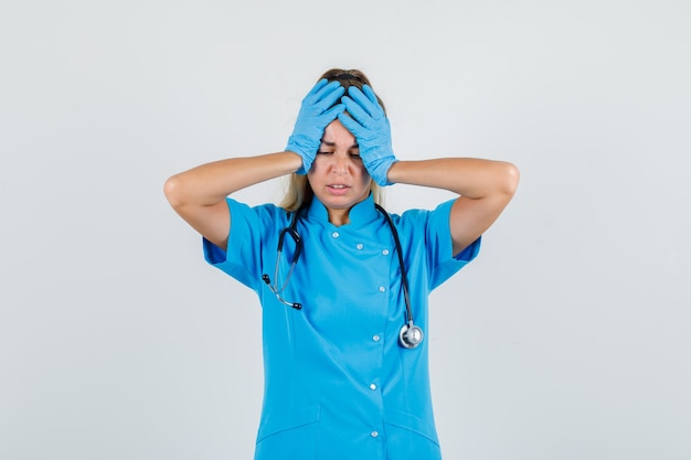 Medico femminile che tiene le mani sulla testa in uniforme blu, guanti e che sembra dispiaciuto.