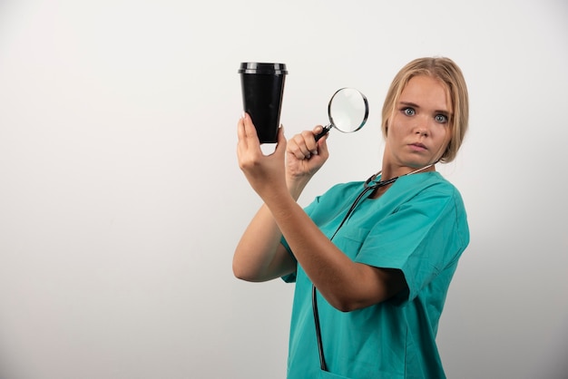 Medico femminile che tiene tazza e lente d'ingrandimento su bianco.