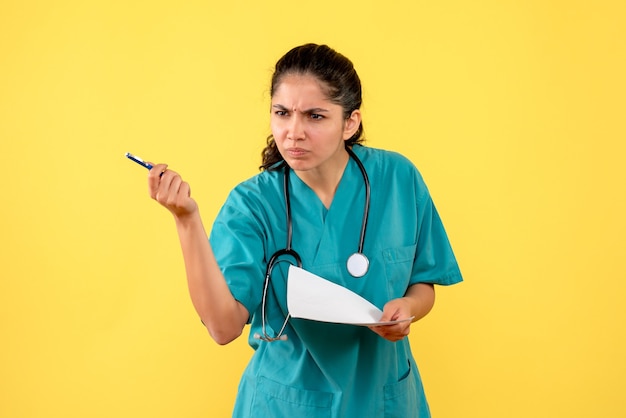 Medico femminile confuso vista frontale che tiene documenti e penna su sfondo giallo