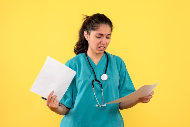 Medico femminile confuso vista frontale con documenti in piedi su sfondo giallo