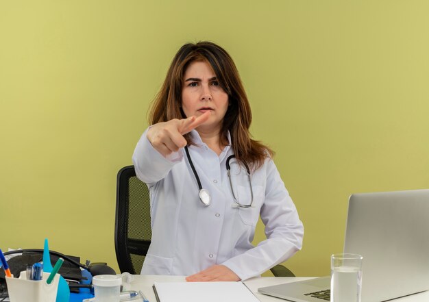 Medico femminile di mezza età accigliato che indossa veste medica e stetoscopio seduto alla scrivania con appunti di strumenti medici e laptop facendo gesto di pistola isolato