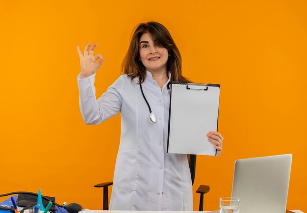 Medico femminile di mezza età che indossa una veste medica con lo stetoscopio in piedi dietro la scrivania lavora sul computer portatile con strumenti medici che tengono appunti e mostra il gesto okey sulla parete arancione con lo spazio della copia