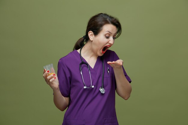 Medico femminile di mezza età concentrato che indossa uniforme e stetoscopio intorno al collo che tiene il contenitore di misurazione con le pillole in esso e la pillola in mano guardando la pillola con la bocca aperta che prende la pillola