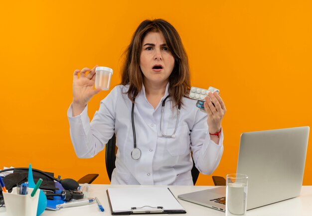 Medico femminile di mezza età confuso che indossa la veste medica con lo stetoscopio che si siede alla scrivania lavora sul computer portatile con strumenti medici che tengono la lattina vuota e le pillole sulla parete arancione isolata