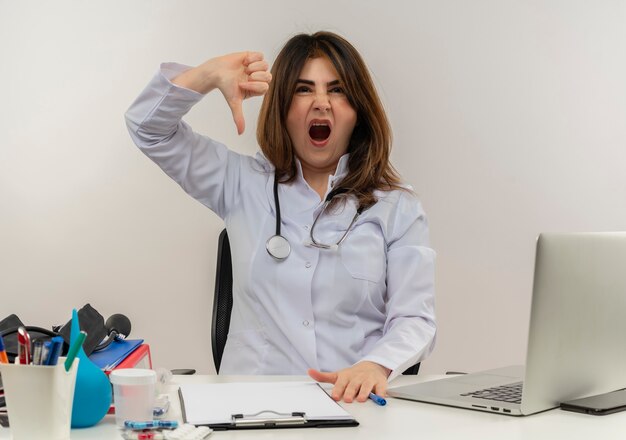 Medico femminile di mezza età dispiaciuto che indossa veste medica e stetoscopio seduto alla scrivania con appunti di strumenti medici e laptop che mostra il pollice verso il basso isolato