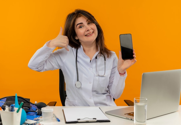 Medico femminile di mezza età gioioso che indossa veste medica e stetoscopio seduto alla scrivania con appunti di strumenti medici e laptop facendo gesto di chiamata che mostra il telefono cellulare isolato